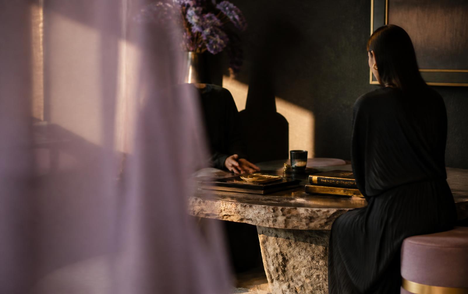 Two people in quiet private conversation across a stone table, sheer lavender fabric drifting in the foreground